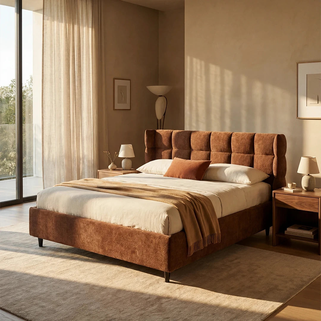 Una camera da letto minimalista inondata di luce ospita il letto imbottito RAGNAR di Letti.store, con contenitore e testiera capitonné avvolgente color marrone, abbinato a biancheria da letto beige e un copriletto color cuoio. Comodini in legno, lampade coordinate, tende trasparenti e opere d’arte dai toni neutri completano il look.
