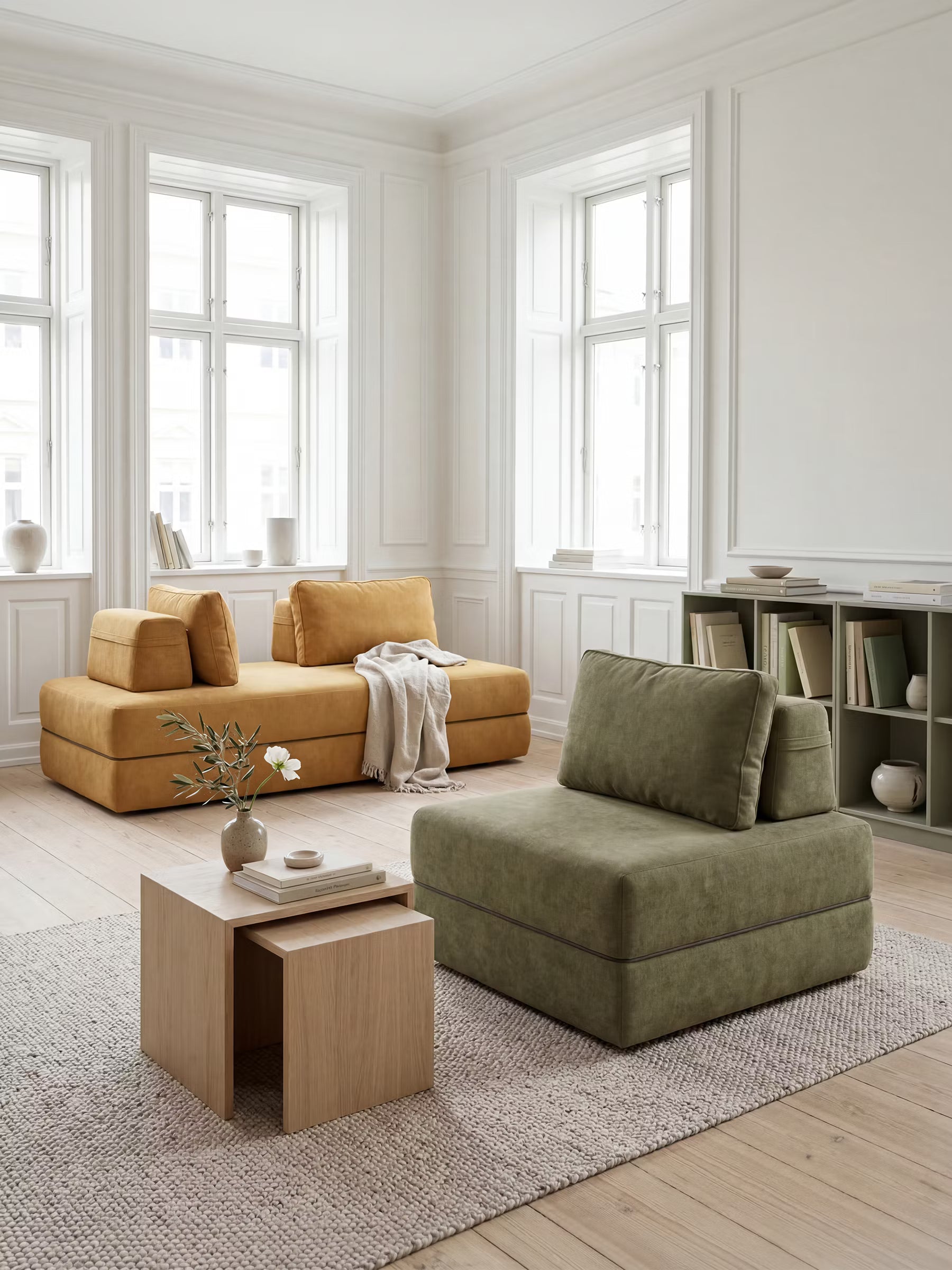 Un soggiorno in stile minimalista con ampie finestre, un divano giallo senape e una poltrona letto Rift di Divani.Store, con materasso alto 24 cm in memory foam, di colore verde oliva, posizionata su un tappeto beige. Due tavolini in legno chiaro, uno dentro l’altro, ospitano un vaso di fiori e una tazza. Una libreria con libri e oggetti decorativi riveste la parete destra, mentre la luce entra a fiotti.