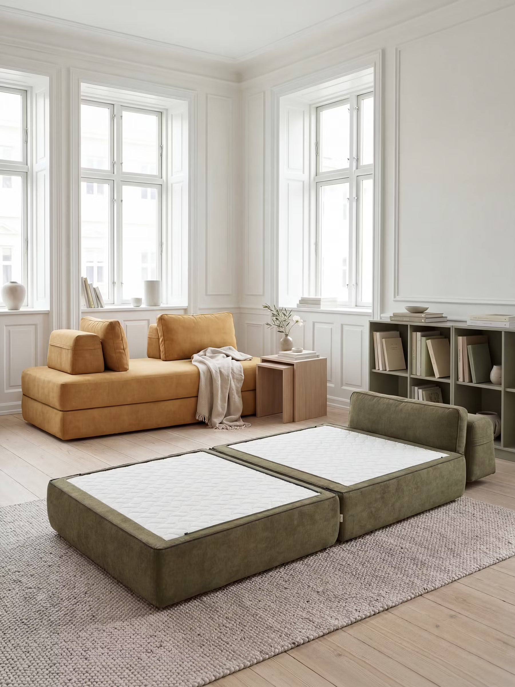 Un soggiorno luminoso e moderno, con ampie finestre, è arredato con un divano componibile giallo senape, un tavolo in legno chiaro e due materassi da pavimento Divani.Store Rift con materassi in memory foam alti 24 cm, schienali regolabili e rivestimento in tessuto T31 facile da pulire, di colore verde oliva, su un tappeto beige testurizzato. Sulla parete di destra, una scaffalatura espone libri e oggetti decorativi.