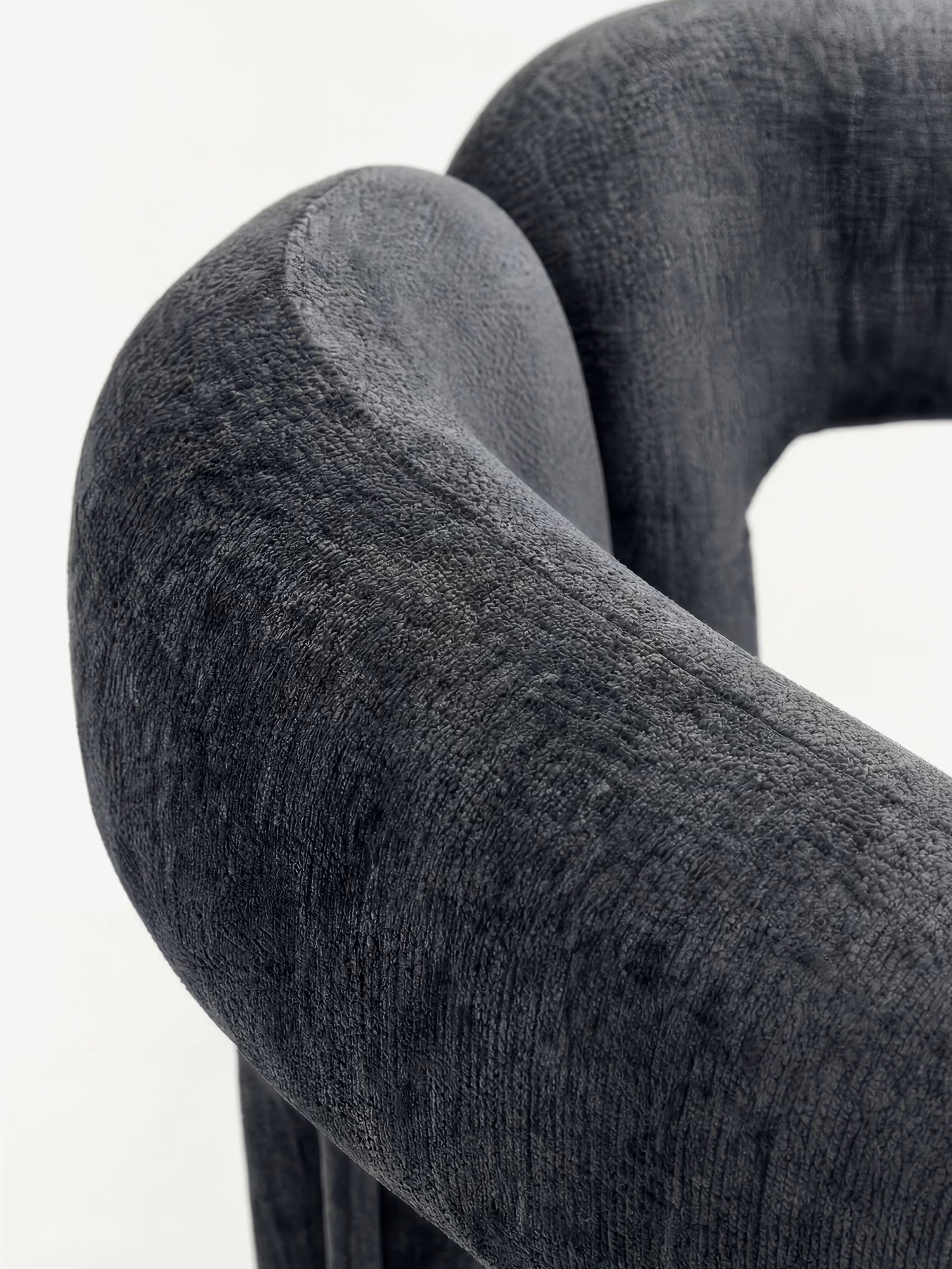 Primo piano della sedia di design imbottita “Sibilla” di Divani.Store, caratterizzata da un morbido tessuto grigio scuro, una forma arcuata interamente imbottita e linee morbide che ne esaltano il comfort contemporaneo su uno sfondo chiaro.