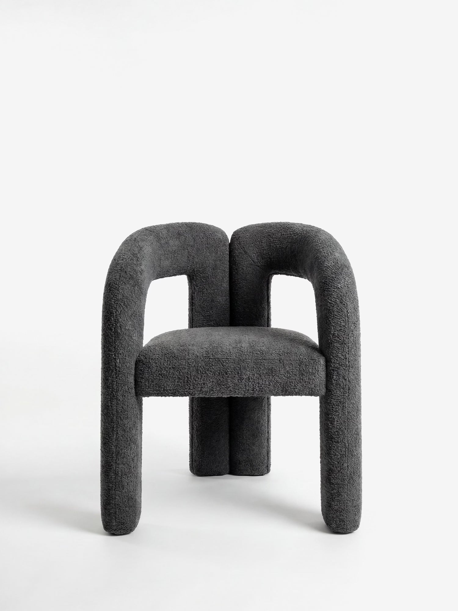 La sedia di design Sibilla di Divani.Store è interamente imbottita in tessuto grigio scuro con struttura rivestita, gambe e braccioli arrotondati e spessi, braccioli ampi e ricurvi, schienale basso e dritto e una forma ad arco scultorea su sfondo bianco.