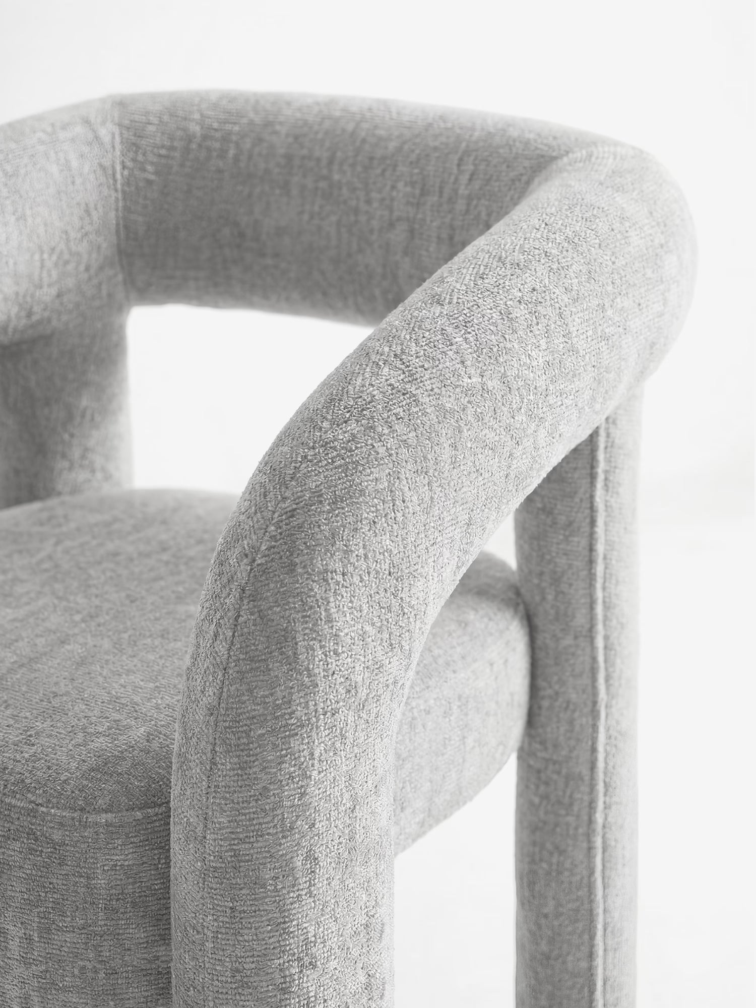 Un primo piano di Sibilla, la sedia di design imbottita di Divani.Store, caratterizzata da un tessuto testurizzato grigio chiaro, una struttura ad arco interamente imbottita, schienale e seduta arrotondati e ben imbottiti, curve morbide e gambe cilindriche su uno sfondo bianco a tinta unita.