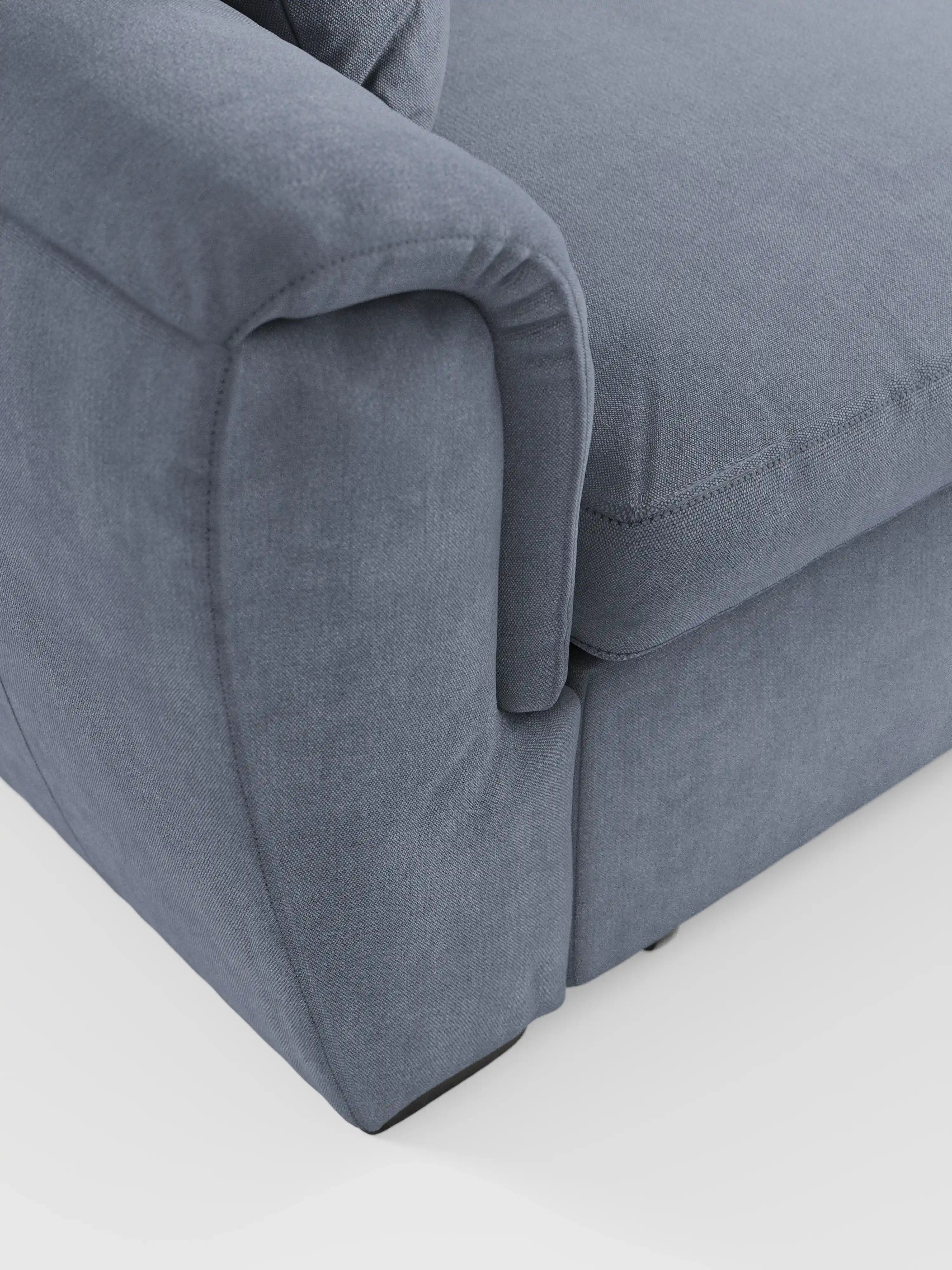 Primo piano del bracciolo e del cuscino di seduta del divano angolare reversibile con contenitore estraibile e pouf di Divani.Store. Rivestito in morbido tessuto Smacchiabile Oeko-Tex® blu-grigio, il divano presenta cuciture a vista lungo i bordi. Il suo design mette in risalto i braccioli arrotondati e le linee morbide e pulite con poggiatesta regolabili, il tutto su uno sfondo bianco a tinta unita.