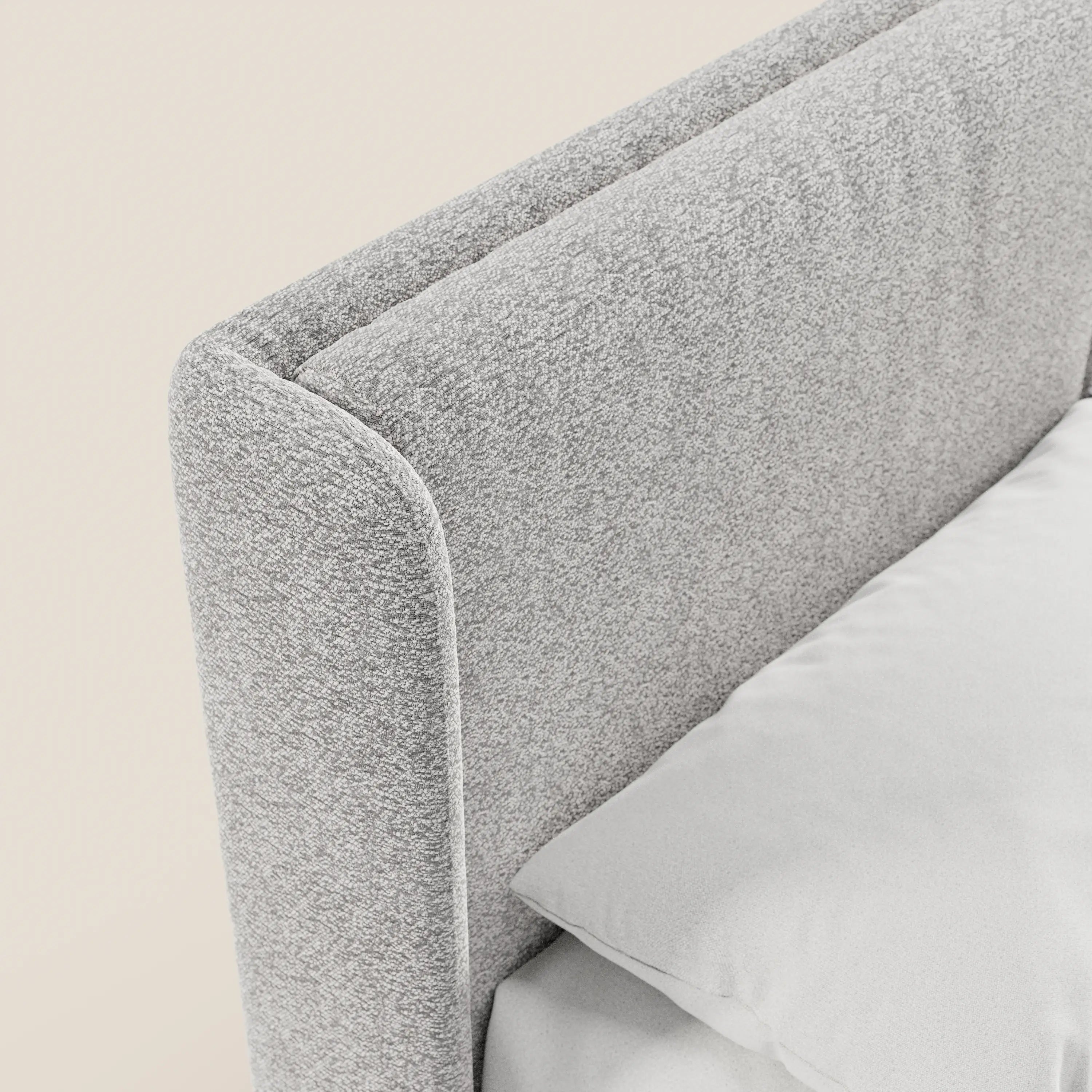 Primo piano del letto di design VALKIRIA di Letti.store, caratterizzato da un tessuto grigio chiaro certificato Oeko-Tex®, una testiera avvolgente con 2 cuscini imbottiti e una base contenitore. La biancheria da letto bianca ricopre il materasso su uno sfondo bianco sporco.