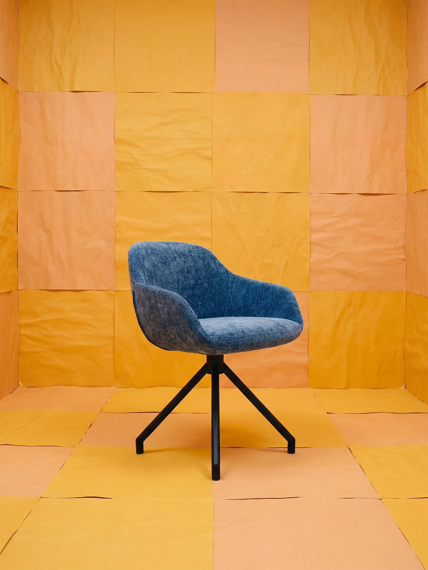 La poltrona imbottita Vico di Divani.Store, realizzata in tessuto soft-touch OEKO-TEX® blu e con gambe in metallo nero, è al centro di una stanza con pareti e pavimento a quadrati arancioni e gialli, creando uno sfondo geometrico minimalista.