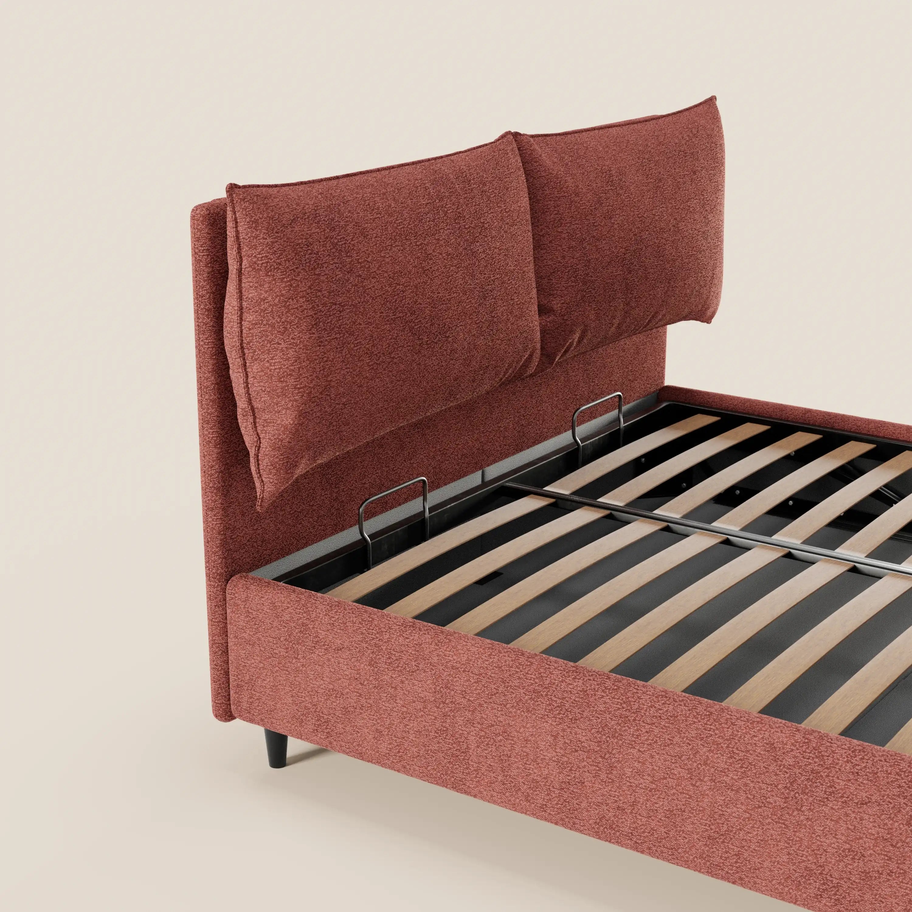 Il letto imbottito ZAYRON di Letti.store presenta una struttura in tessuto color ruggine certificato Oeko-Tex®, due grandi cuscini regolabili che fungono da testiera, doghe in legno a vista e supporti in metallo nero. In questa immagine è visibile solo una parte di questo moderno letto contenitore, su uno sfondo beige chiaro.