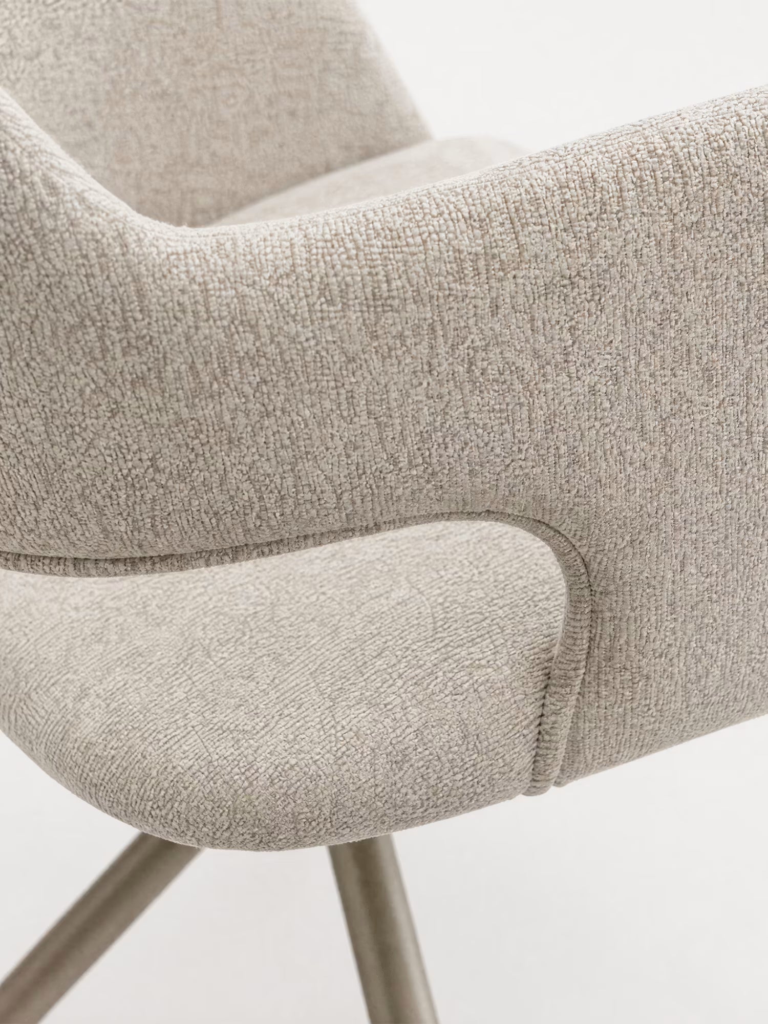 Primo piano della sedia imbottita di design Zefira di Divani.Store, caratterizzata da un tessuto effetto cotone in beige chiaro, braccioli ricurvi, cuciture discrete e una base in metallo color champagne su sfondo bianco.