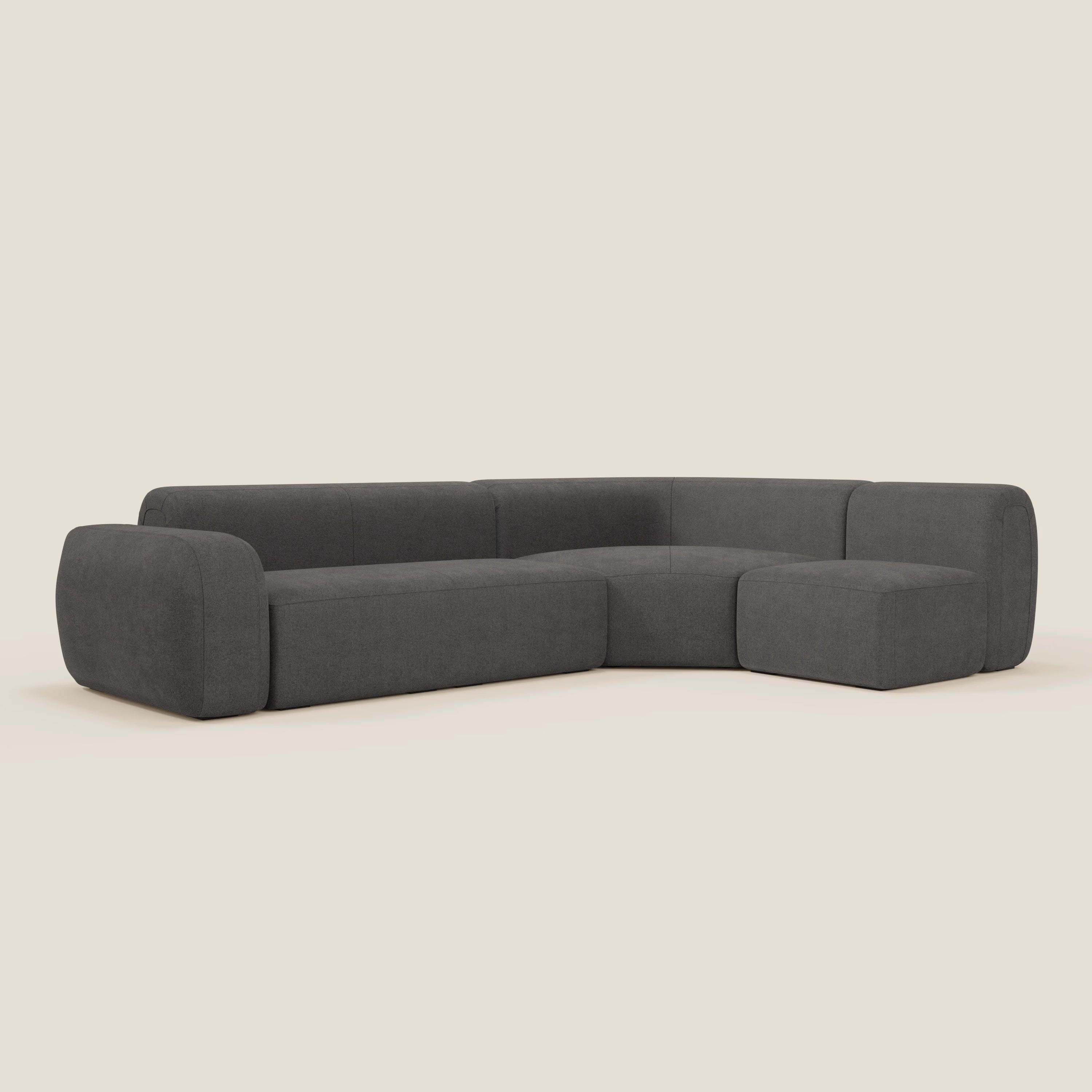 Il divano angolare componibile e modulare BonBon di Divani.Store, in tessuto smacchiabile T23 antracite, è caratterizzato da un design minimalista con braccioli e schienali arrotondati, sedute basse e larghe e cuscini spessi su uno sfondo bianco sporco.