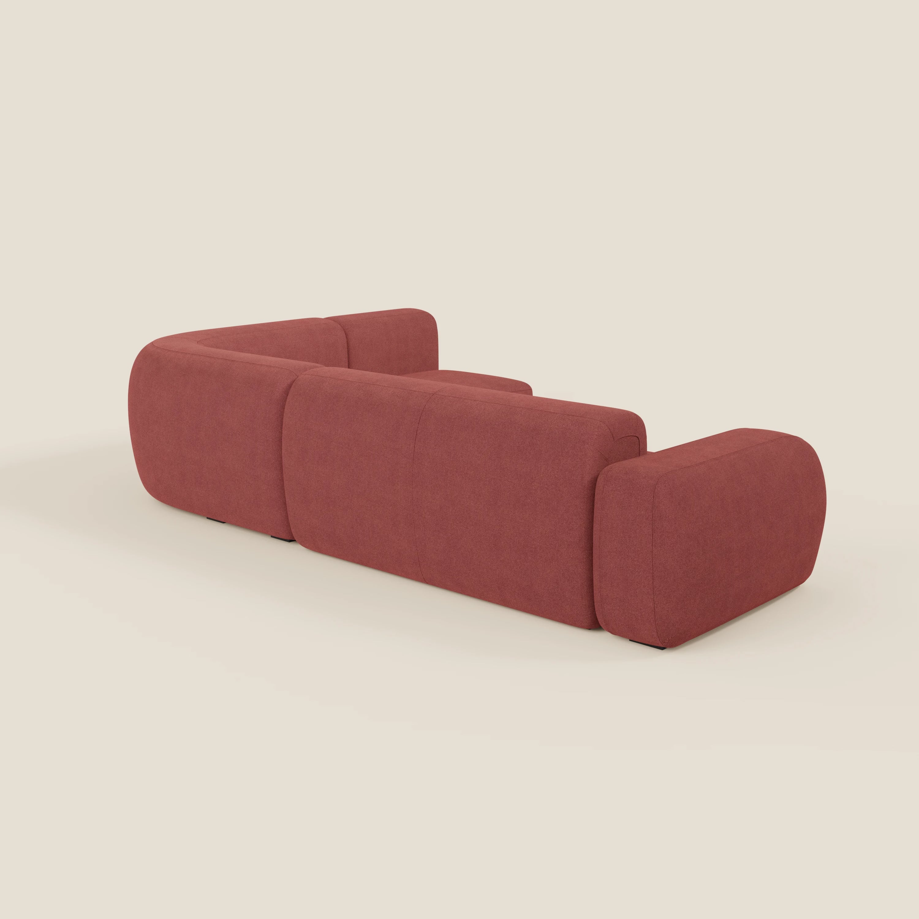 Il divano BonBon angolare componibile e modulare di Divani.Store è caratterizzato da bordi arrotondati e braccioli spessi e imbottiti in tessuto antimacchia T23 rosso ciliegia. Mostrato dal retro su uno sfondo beige chiaro. Prodotto in Italia.