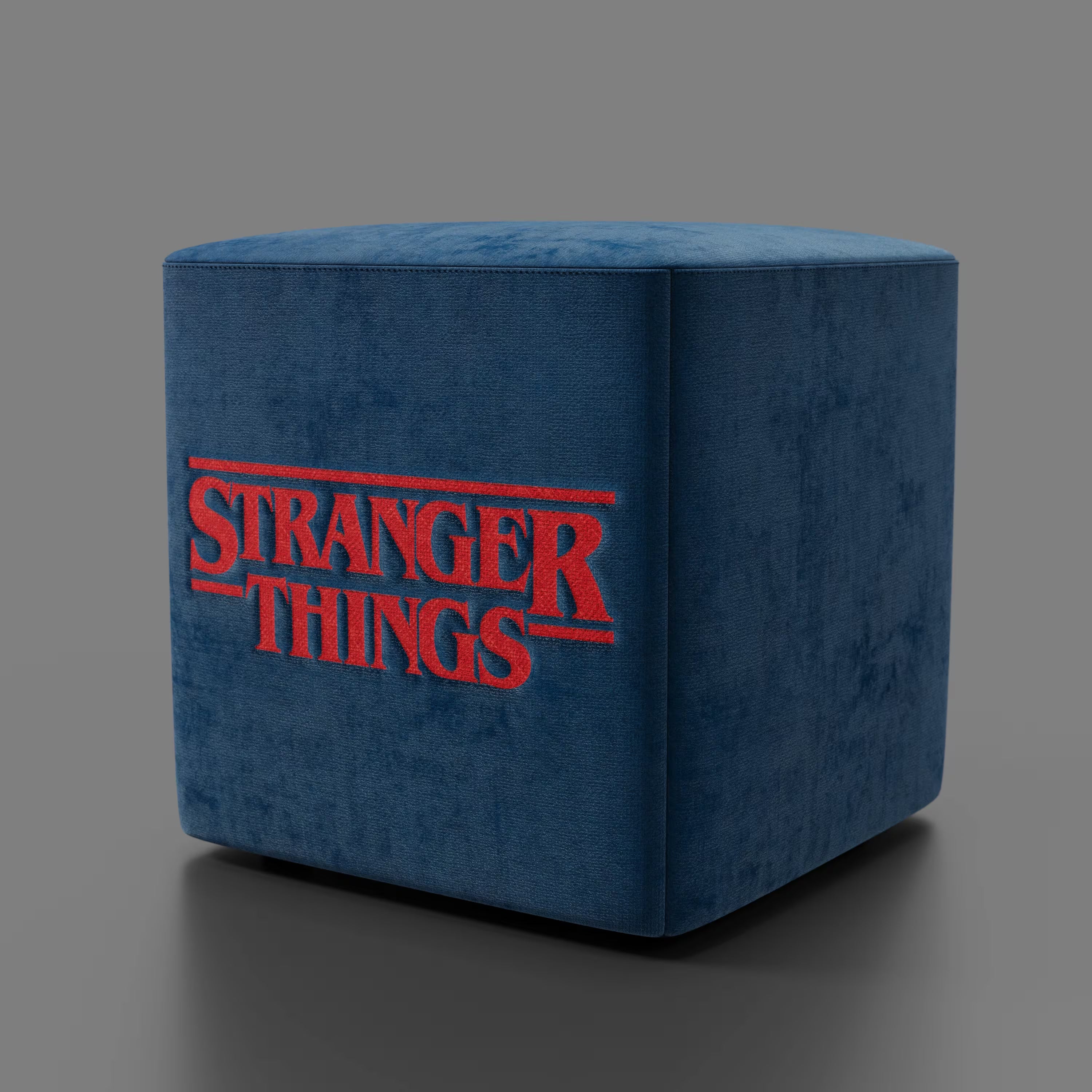 Il pouf Cube Stranger Things Edition di Divani.Store è caratterizzato da un tessuto blu antimacchia, una texture simile al velluto e il logo rosso della serie su un lato, su uno sfondo grigio.