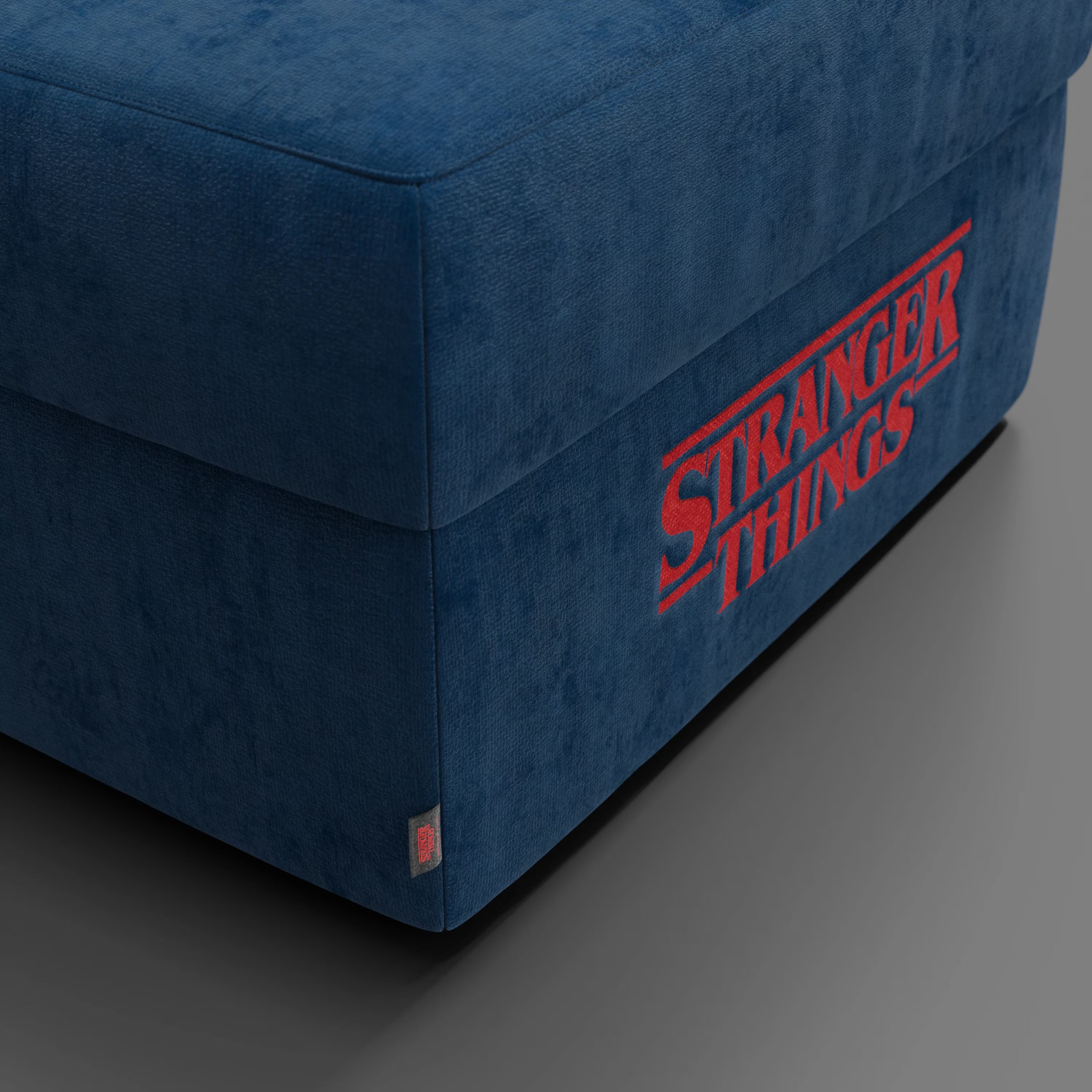 Primo piano del pouf Divani.Store Sun Contenitore Stranger Things Edition (60x80 cm, tessuto smacchiabile blu) con la scritta Netflix in rosso, su un pavimento grigio. È visibile una piccola etichetta in tessuto, perfetta per qualsiasi arredamento Netflix.