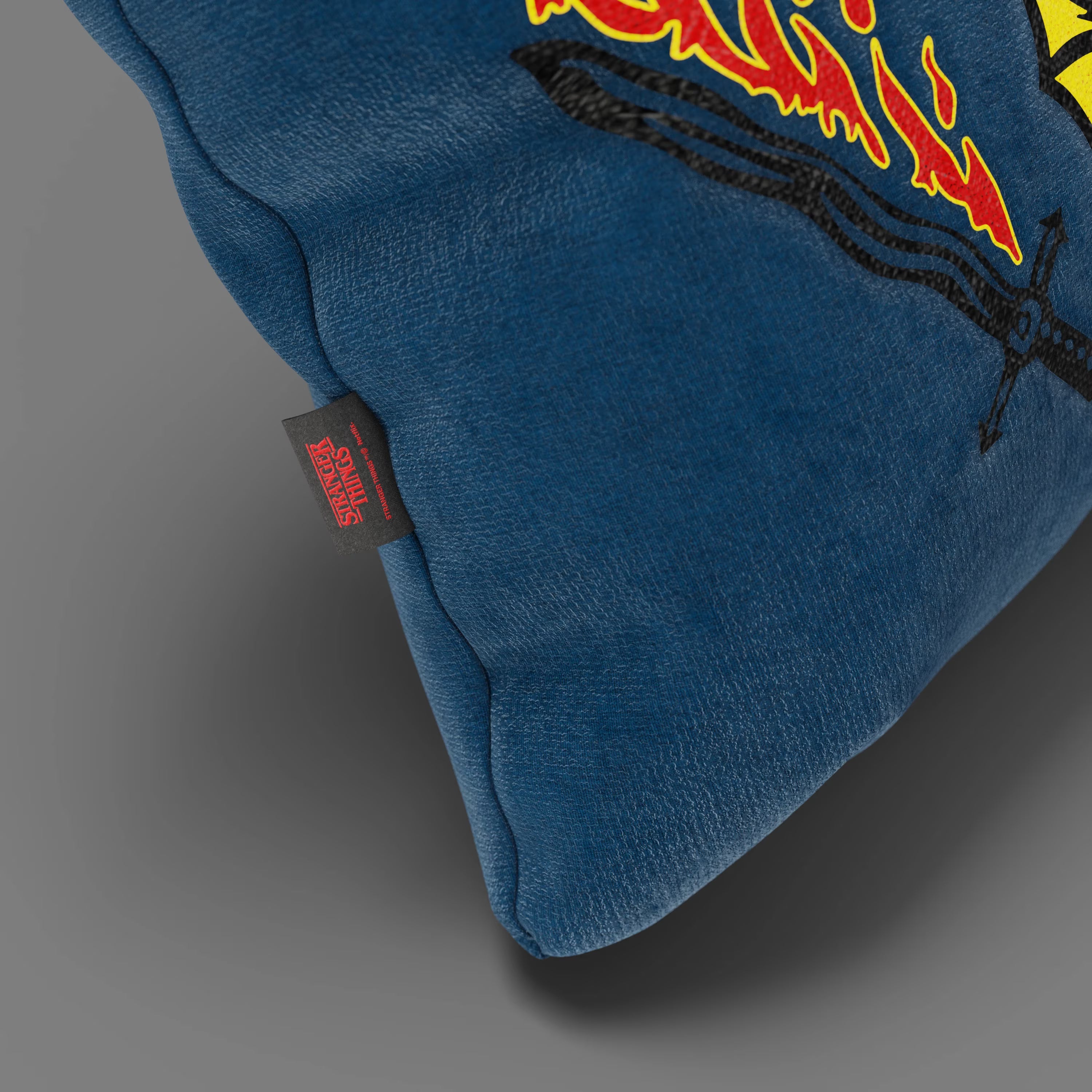 Primo piano del Cuscino Hellfire Club Stranger Things Limited Edition di Divani.Store in blu (45x45), con grafica ricamata di fiamme e spade, targhetta nera con scritta rossa "Stranger Things", su sfondo grigio.