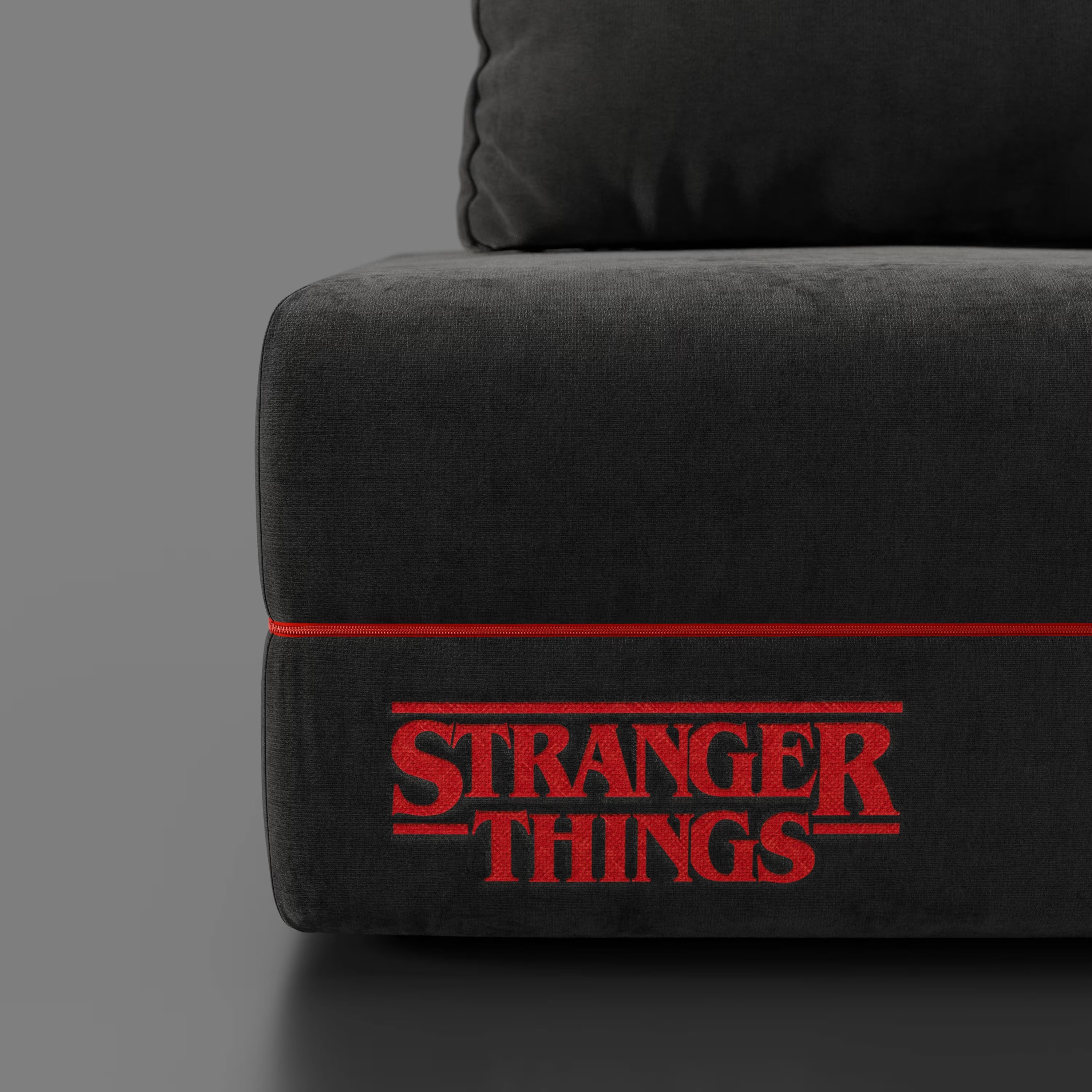 Un primo piano della poltrona letto Divani.Store Rift STRANGER THINGS Edition in smacchiabile antracite, con profili rossi e logo sul davanti. Lo sfondo grigio chiaro mette in risalto il materasso in memory foam da 22 cm e i dettagli di design unici.