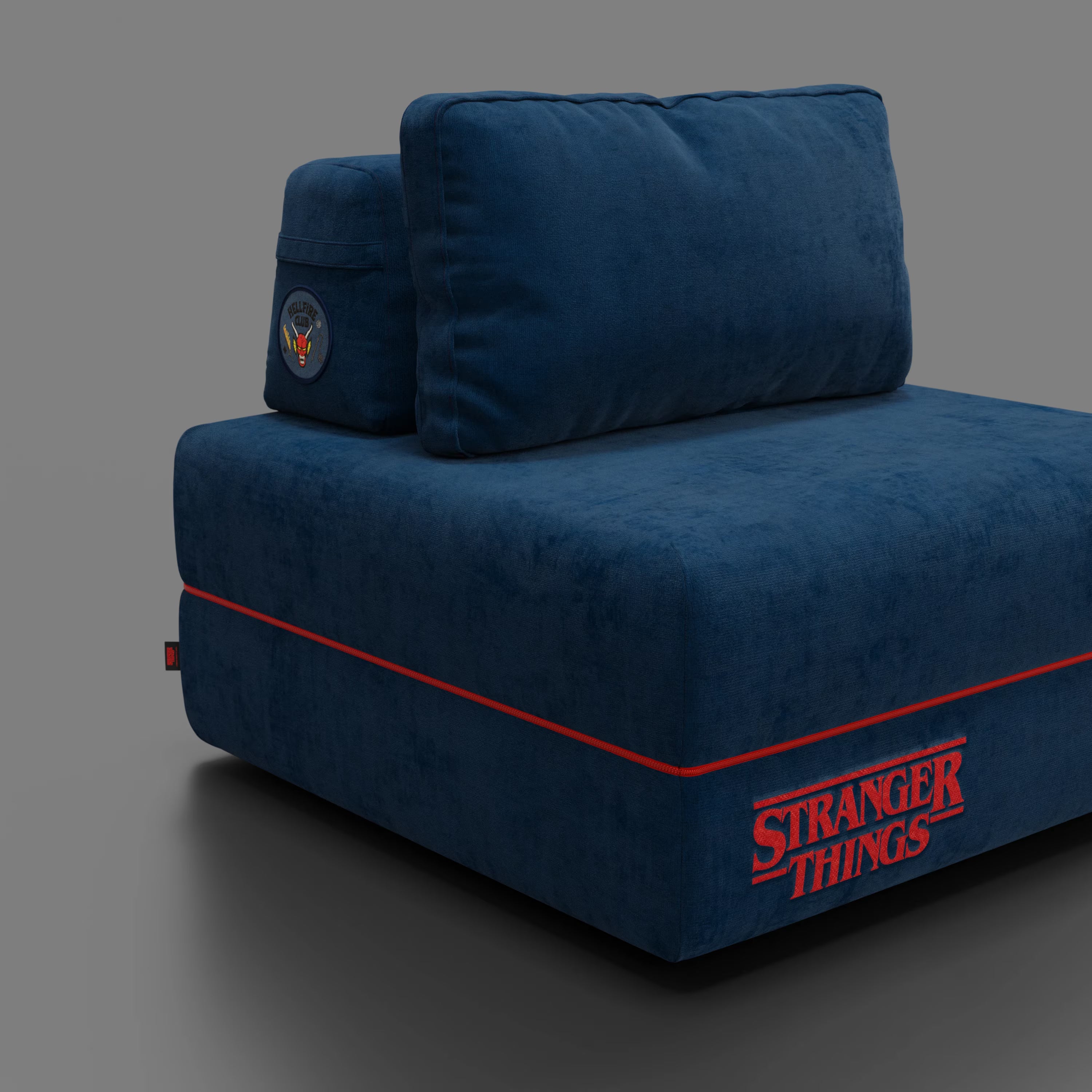 Una poltrona letto Rift blu navy STRANGER THINGS Edition di Divani.Store, con profili rossi, logo "Stranger Things" ricamato in rosso e patch del laboratorio Hawkins sul poggiatesta, si trova su uno sfondo grigio.
