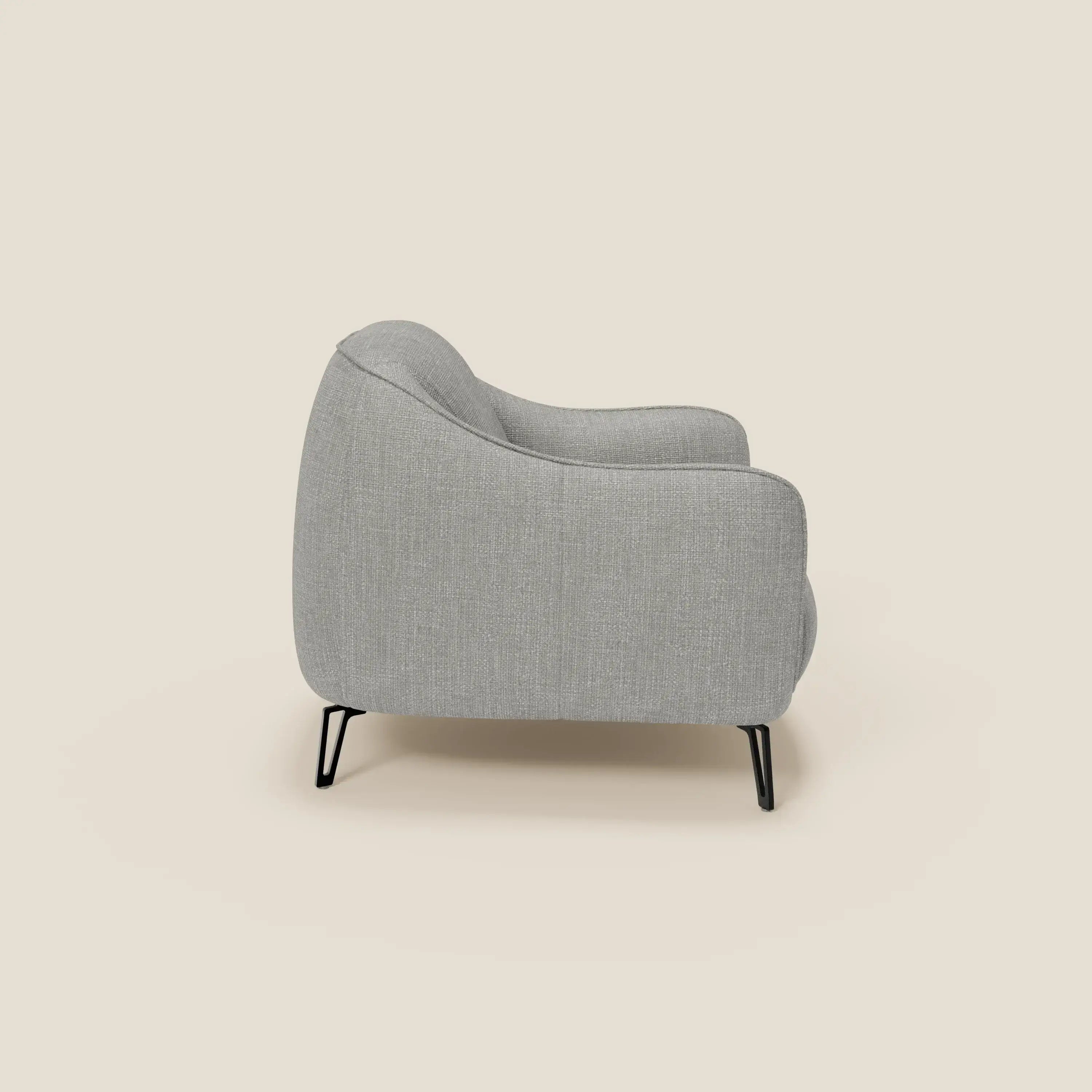 La poltroncina Avela di Divani.Store, raffigurata di profilo, è caratterizzata da schienale e braccioli curvi, eleganti gambe in metallo nero e rivestimento in morbido tessuto impermeabile T06 grigio su sfondo beige.
